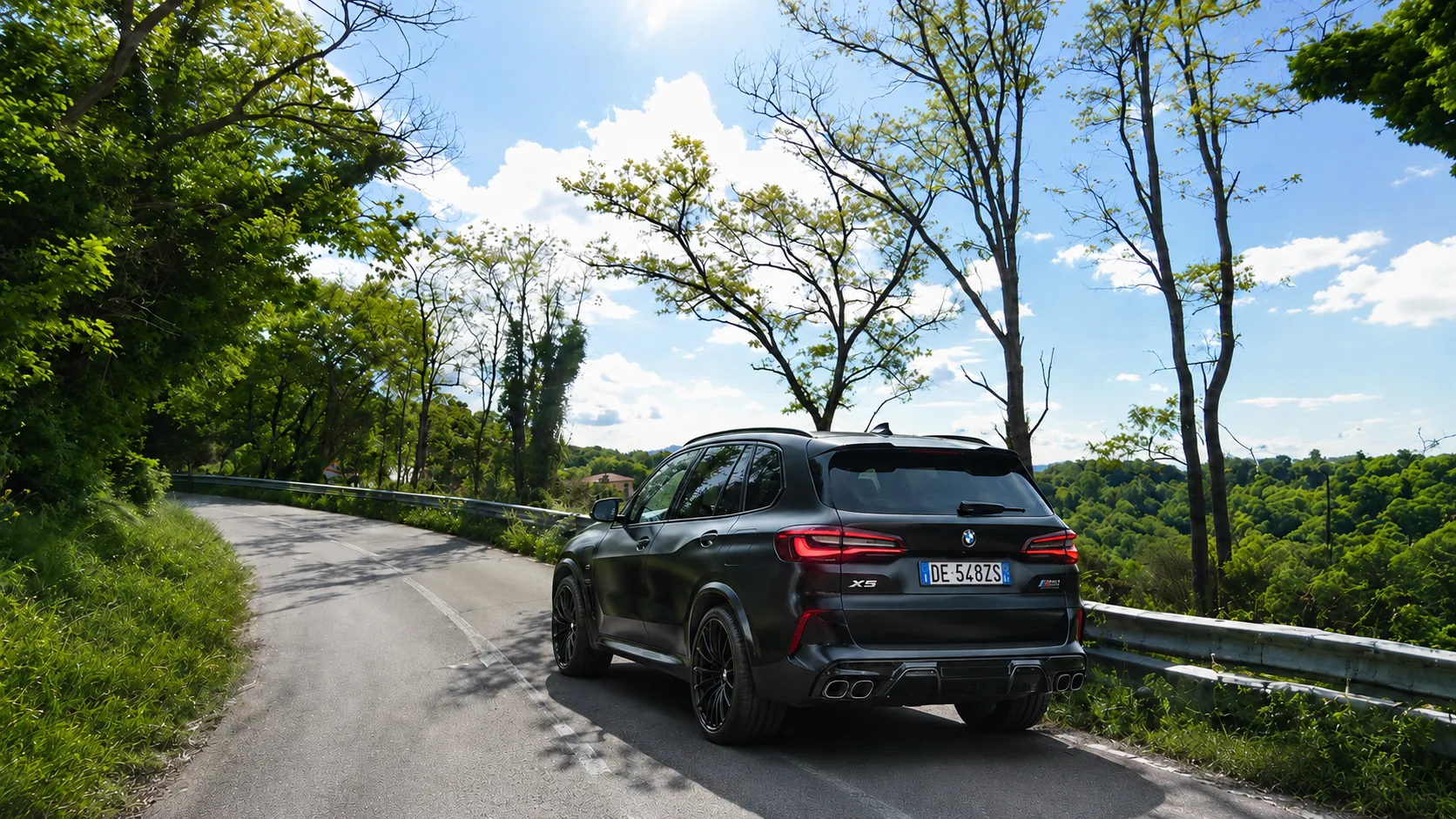 A car winds along a tree-lined hillside road above the green valley